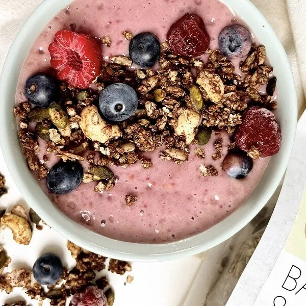 Smoothie Bowl mit rosa Joghurt, Pure&Sweet granola, Blaubeeren und Himbeeren in einer weißen Schüssel.