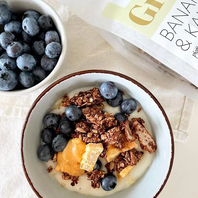 Schüssel mit Joghurt, Pure&Sweet granola, Blaubeeren und Honigstücken; daneben eine Schüssel Blaubeeren und eine Packung granola.