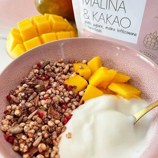 Rosa Schüssel mit Pure&Sweet Malina & Kakao granola, Joghurt und Mangostücken; im Hintergrund die Produktverpackung und eine Mangofrucht.
