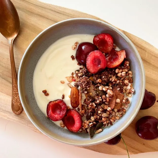Schüssel mit Joghurt, Pure&Sweet granola, frischen Kirschen und Mandeln auf einem Holzbrett, daneben ein kupferner Löffel.