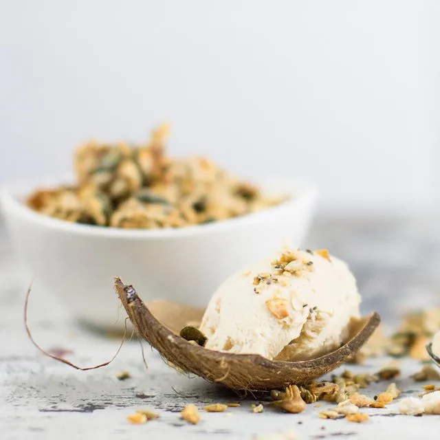 Kugel Kokosnusseis in einer Kokosnusshälfte, bestreut mit Pure&Sweet granola; im Hintergrund eine weiße Schüssel mit granola.